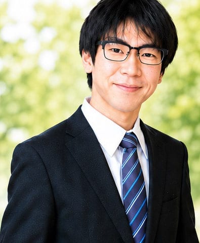 ライフサポート熊谷 代表の齊藤匠。緑の木々を背景に、スーツ姿で微笑んでいる